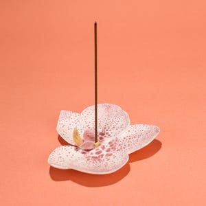May include: An orchid-shaped incense holder in white and pink, with a brown incense stick. The holder features pink spots and a yellow centre. The background is a solid coral colour. The incense stick is upright.