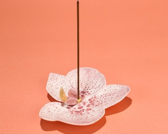 May include: A white and pink orchid-shaped incense holder with a brown incense stick. The holder has pink spots and a yellow center. The background is a solid coral color. The incense stick is standing upright.