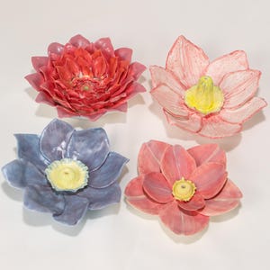 Pode incluir: Quatro esculturas de flores de cerâmica em tons de vermelho, rosa e azul. Cada flor tem um centro amarelo. As flores estão dispostas sobre uma superfície branca. As flores são provavelmente itens decorativos.