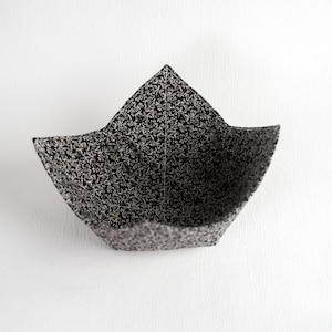 May include: A black and white patterned fabric bowl, shaped like a boat or a crown, sits against a white background. The bowl's interior and exterior feature a repeating floral design. The fabric is folded and stitched to create the bowl's form.