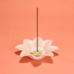 Peut inclure: Un porte-encens en céramique rose clair en forme de fleur, avec un centre vert. Un bâton d'encens marron est inséré au centre. Le fond est de couleur pêche.