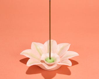 May include: A light pink ceramic incense holder shaped like a flower with a green center. A brown incense stick is inserted in the center of the flower. The background is a solid peach color.