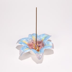 Op de afbeelding: Een keramische wierookhouder in de vorm van een leliebloem. De bloem is beschilderd in blauwe, roze en gele tinten. Een bruine wierookstok zit in het midden van de bloem, die op een witte basis staat.