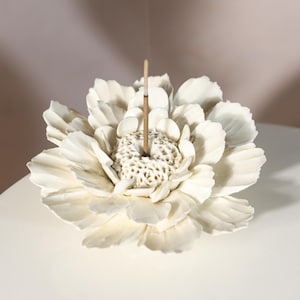 Puede incluir: Un portaincienso de cerámica blanca con forma de flor de peonía. La flor tiene pétalos detallados y un centro texturizado. Un palito de incienso está encendido y emitiendo humo. El soporte está sobre una superficie blanca.