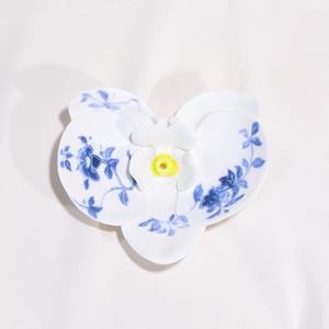Puede incluir: Un plato blanco de cerámica con forma de flor con motivos florales azules y un centro amarillo. El plato tiene un diseño tridimensional, con pétalos que se curvan hacia arriba. El patrón floral azul es detallado y delicado.