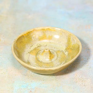 May include: A ceramic incense holder with a raised center cone. The dish is round with a speckled, glazed finish in shades of beige, yellow, and gold. The incense holder is designed to catch ash.