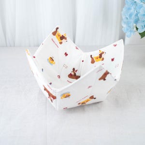 May include: White fabric bowl with a repeating pattern of brown dogs wearing yellow coats, coffee cups, and red butterflies. The words "Happy winter" are printed on the fabric. The bowl is folded into a square shape.