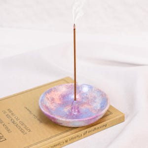 Puede incluir: Un porta-incienso de cerámica redondo y poco profundo con un acabado moteado en tonos pastel de rosa, azul y morado. Una varilla de incienso marrón está insertada en el orificio central, emitiendo un fino hilo de humo blanco al aire.
