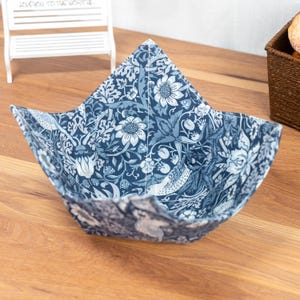 May include: A navy blue fabric bowl featuring a floral and bird pattern in white and blue hues. The bowl is open and square-shaped, designed for food. The fabric has a detailed, vintage-inspired design, perfect for serving.