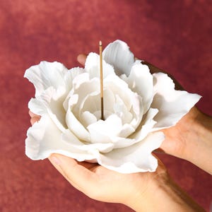 May include: A white, flower-shaped incense holder with a single incense stick. The holder is held in two hands against a burgundy background. The flower petals are detailed and layered, creating a realistic appearance.