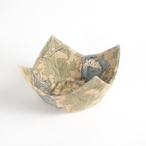 Puede incluir: Un cuenco de tela con un estampado floral en tonos verdes, azules y beige. El cuenco es abierto y tiene forma cuadrada con esquinas dobladas. La tela parece ser de algodón o un material similar.