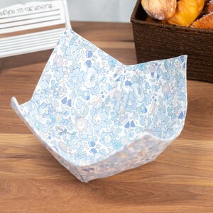 Puede incluir: Un cuenco de tela con un estampado floral en tonos azules y blancos. El cuenco está abierto y se encuentra sobre una superficie de madera. El cuenco está diseñado para contener alimentos.