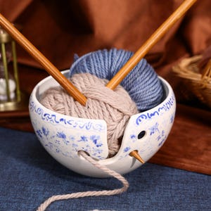 Puede incluir: Un cuenco de cerámica blanca para lana con un patrón floral azul, que contiene un ovillo de lana beige y uno de lana azul. Dos agujas de tejer de madera están insertadas en el cuenco, con una hebra de lana beige saliendo por la ranura lateral.