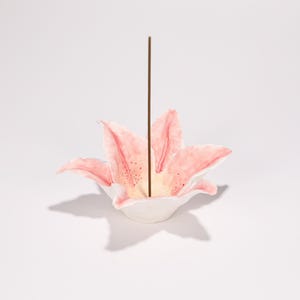 May include: A ceramic incense holder in the shape of a pink and white lily flower. A brown incense stick is placed in the centre. The lily petals have pink watercolor shading and small brown spots.