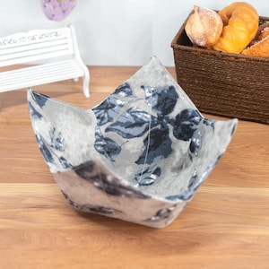 Op de afbeelding: Een grijze stoffen kom met een bloemenpatroon in blauwe en grijze tinten. De kom heeft een vierkante vorm met gevouwen hoeken en witte stiksels. Hij staat op een houten oppervlak.