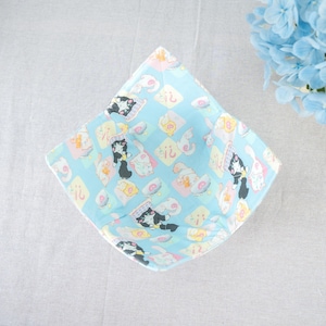 Może przedstawiać: Jasnoniebieska miska z tkaniny z powtarzającym się wzorem kreskówkowych kotów, filiżanek do herbaty i innych elementów dekoracyjnych. Miska ma kwadratowy kształt z zaokrąglonymi rogami i kontrastującym różowym obramowaniem. Projekt zawiera pastelowe kolory.