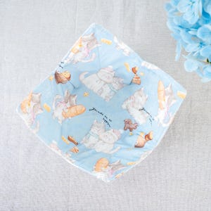 May include: A light blue fabric bowl cozy with a repeating pattern of cartoon cats, bread, and pastries. The text "You make me so happy" is written on the fabric. The cozy is square-shaped and has a white border.