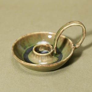 May include: A ceramic candle holder with a circular base and a handle. The candle holder is glazed in green and brown tones, with a dark blue ring around the candle cup. The handle curves above the base. The candle holder is handmade.