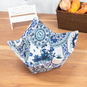 May include: A square, fabric bowl with a blue and white floral pattern. The bowl is open and sits on a wooden surface. The pattern includes circular and floral designs. The bowl is empty.