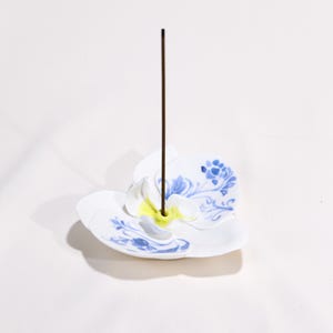 Puede incluir: Porta incienso de cerámica blanca con forma de flor con diseños florales azules. Un palito de incienso marrón se alza en el centro. El soporte tiene un centro amarillo y está sobre un fondo blanco.