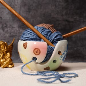 May include: A decorative ceramic yarn bowl holding blue and brown yarn. The bowl is white with pink, brown, and blue patterns, and wooden knitting needles are inserted. A blue yarn strand trails from the bowl.