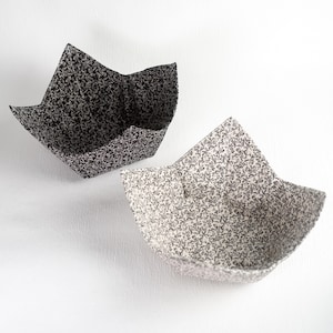 Puede incluir: Dos cuencos de tela con estampado floral. Uno es blanco y negro, el otro blanquecino con un diseño floral similar en blanco y negro. Los cuencos están doblados en forma cuadrada con cuatro lados hacia arriba.