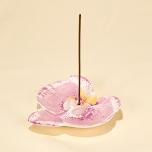Könnte beinhalten: Ein rosa-weißer Keramik-Räucherstäbchenhalter in Form einer Orchideenblüte. Ein brauner Räucherstäbchen-Stick steht in der Mitte. Der Halter hat ein gesprenkeltes Design und gelbe Akzente.