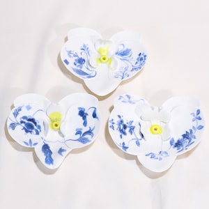 Puede incluir: Tres platos blancos de cerámica con forma de orquídea, con diseños florales azules y centros amarillos. Los platos están dispuestos sobre una superficie blanca, mostrando el delicado diseño. Los patrones azules varían ligeramente.