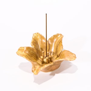 Puede incluir: Un porta incienso con forma de flor de color dorado. El soporte de cerámica tiene pétalos detallados y un área central para palitos de incienso. Un solo palito de incienso marrón está insertado en el centro.