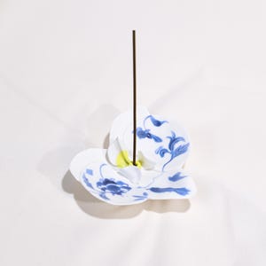 Puede incluir: Porta incienso de cerámica blanca con diseños florales azules. El soporte tiene forma de flor con una varilla de incienso marrón en el centro. Presenta un acento amarillo.