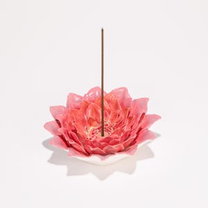 May include: A ceramic incense holder shaped like a pink lotus flower with a brown incense stick. The flower has multiple layers of petals and sits on a white base. The incense stick is centered in the flower.