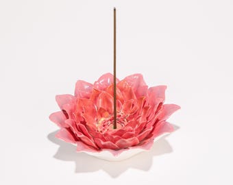 May include: A ceramic incense holder shaped like a pink lotus flower with a brown incense stick. The flower has multiple layers of petals and sits on a white base. The incense stick is centered in the flower.