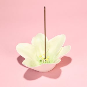 Könnte beinhalten: Ein weißer Keramik-Räucherstäbchenhalter in Form einer Blume mit einem braunen Räucherstäbchen. Die Blume hat eine hellgrüne Mitte mit gelben Akzenten. Der Halter steht auf einer rosa Oberfläche.