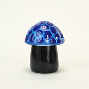 Puede incluir: Una figura decorativa de hongo con una tapa azul y azul claro estampada y un tallo negro. La seta es una pieza de acento caprichosa, probablemente de cerámica o madera pintada, y se encuentra sobre una superficie blanca.