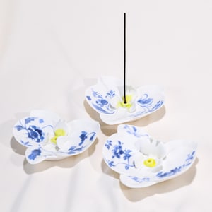 Puede incluir: Tres porta inciensos de cerámica blanca con forma de flores de orquídea, con diseños florales azules. Un soporte tiene un palito de incienso encendido. Los soportes tienen un centro amarillo y están colocados sobre una superficie blanca.