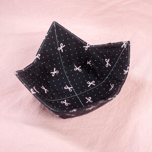 Puede incluir: Un protector de cuenco negro con un estampado de lunares blancos y lazos. El protector de cuenco tiene forma cuadrada con esquinas y bordes cosidos. El interior es negro con un estampado de lunares blancos y lazos. Diseñado para sujetar un cuenco de comida.