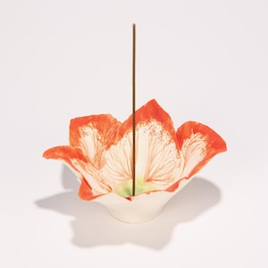 May include: A ceramic incense holder shaped like a flower with orange and white petals. A brown incense stick stands upright in the center. The holder has a white base and a green center.