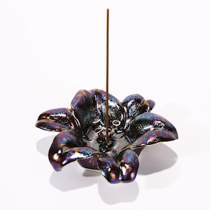 Pode incluir: Um porta-incenso de cerâmica em forma de flor com tons iridescentes roxos, azuis e dourados. Um palito de incenso castanho está no centro. O prato em forma de flor tem várias pétalas e um acabamento brilhante.