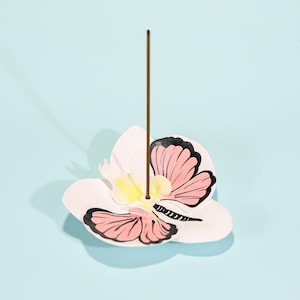Könnte beinhalten: Weißer Keramik-Räucherstäbchenhalter in Form einer Blume mit Schmetterlingsdesign. Der Schmetterling hat rosa Flügel mit schwarzen Umrissen. Ein brauner Räucherstäbchen-Stick ist in der Mitte eingesetzt. Der Hintergrund ist hellblau.