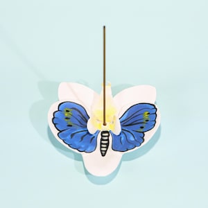Puede incluir: Un porta incienso de cerámica blanca con forma de flor, con un diseño de mariposa azul con detalles negros y acentos amarillos. Un palito de incienso marrón está insertado en el soporte, sobre un fondo azul claro.