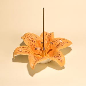 Könnte beinhalten: Orangefarbener, lilienförmiger Räucherstäbchenhalter mit dunkelvioletten Punkten und einem braunen Räucherstäbchen. Die Keramikblume hat einen weißen Sockel und steht vor einem hellgelben Hintergrund. Dekorativer Artikel.