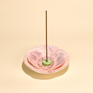 Puede incluir: Un porta incienso de cerámica rosa con forma de flor con una varilla de incienso marrón. El soporte tiene un centro verde y un diseño moteado. El fondo es de color beige claro liso.