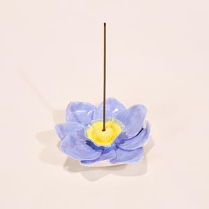 Könnte beinhalten: Ein Keramik-Räucherstäbchenhalter in Form einer blauen Lotusblume mit gelbem Zentrum. Ein brauner Räucherstäbchen ist in der Mitte der Blume eingesetzt. Der Halter ist ein dekorativer Artikel für Aromatherapie und Heimdekoration.