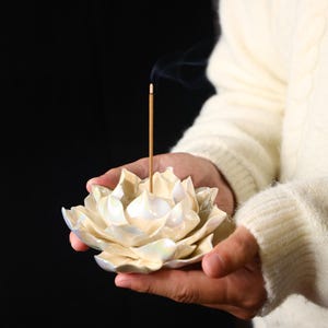 Puede incluir: Un porta incienso de cerámica nacarada con forma de flor de loto con una varilla de incienso encendida. El soporte se sostiene frente a un fondo oscuro. La varilla de incienso emite una fina corriente de humo.