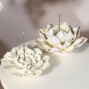 Puede incluir: Dos porta inciensos de cerámica blanca con forma de flor. Cada soporte tiene un pozo central para sostener el palito de incienso. Los soportes están sobre una superficie blanca. Los palitos de incienso son marrones.