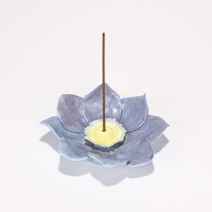May include: A ceramic incense holder shaped like a blue and purple flower with a yellow center. A brown incense stick stands upright in the center. The holder is on a white background.