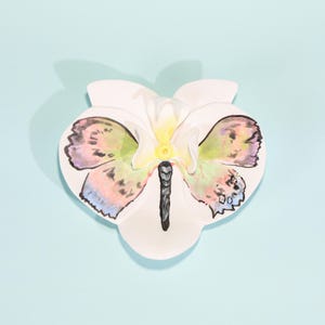 May include: A white ceramic butterfly-shaped ornament with pastel-coloured watercolour wings. The butterfly has a black body, set against a light blue background.