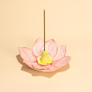 Könnte beinhalten: Ein rosa Lotusblüten-förmiger Räucherstäbchenhalter mit gelbem Zentrum. Ein brauner Räucherstäbchen-Stick steht aufrecht in der Mitte des Halters. Der Halter ist aus Keramik und hat ein handgemaltes Aussehen.