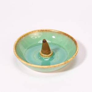 May include: A teal ceramic incense cone burner with a brown rim and a brown incense cone in the center. The burner has a shallow, round design with a glossy finish. The overall aesthetic is minimalist and zen-like.