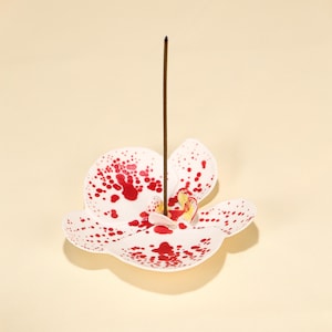 May include: White ceramic incense holder in the shape of an orchid flower, decorated with red speckled patterns. A brown incense stick is placed in the centre. The holder has yellow and red accents in the middle.